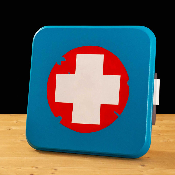 A TF2 Medium Health Kit Replica Props for cosplay featuring its blue lid with a white cross on a chipped paint red circle, the white body and its grey handles.